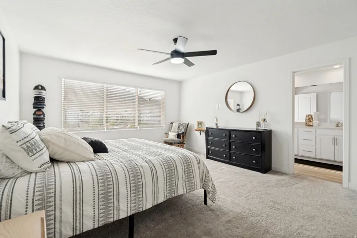 Carpeted bedroom featuring a ceiling fan with integrated lighting, a large window with horizontal blinds, and a doorway opening to a bathroom with a dual vanity