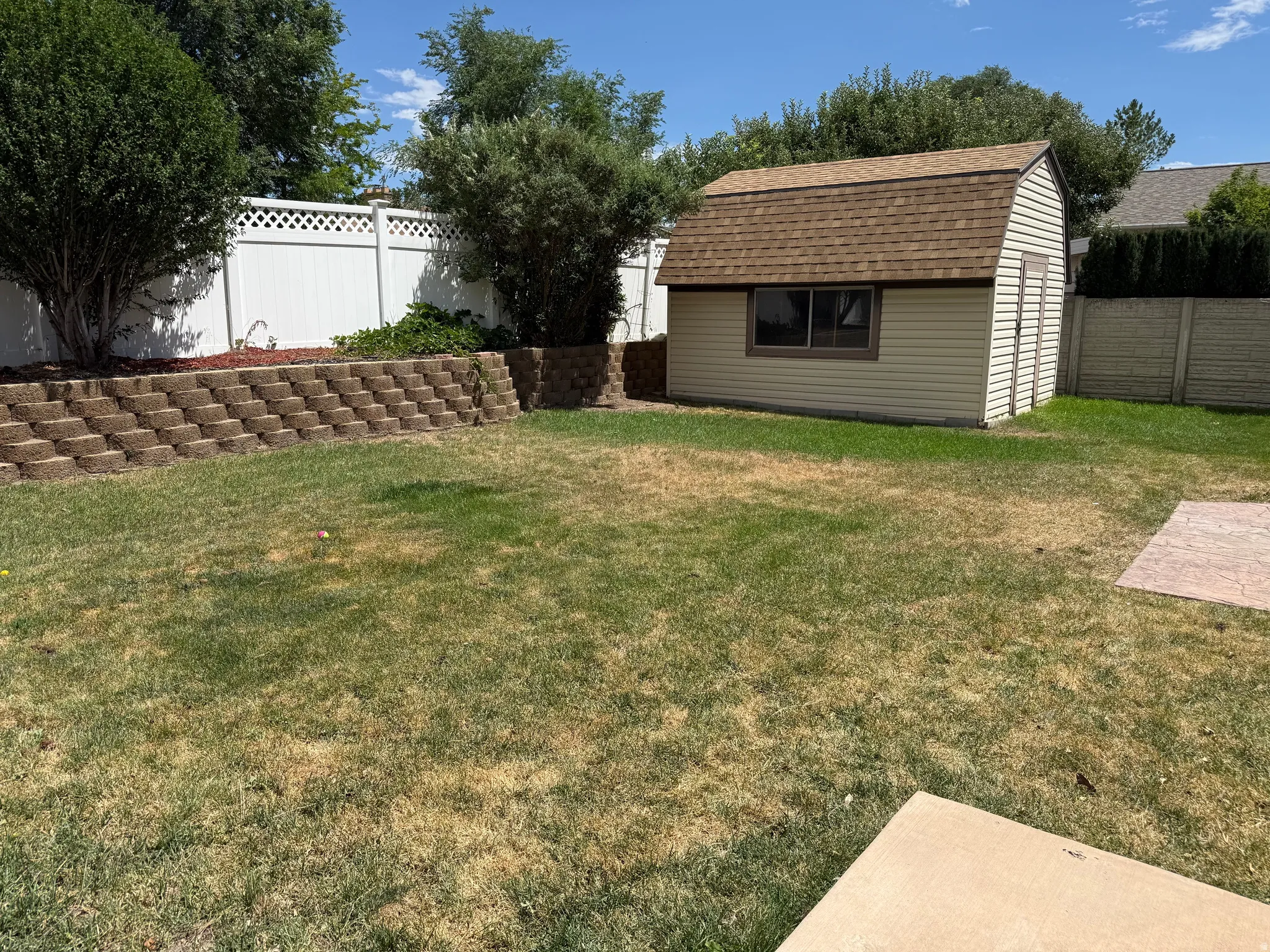 Spacious backyard featuring a utility shed with a gambrel roof and vinyl siding, a white lattice-top privacy fence, and a tiered retaining wall