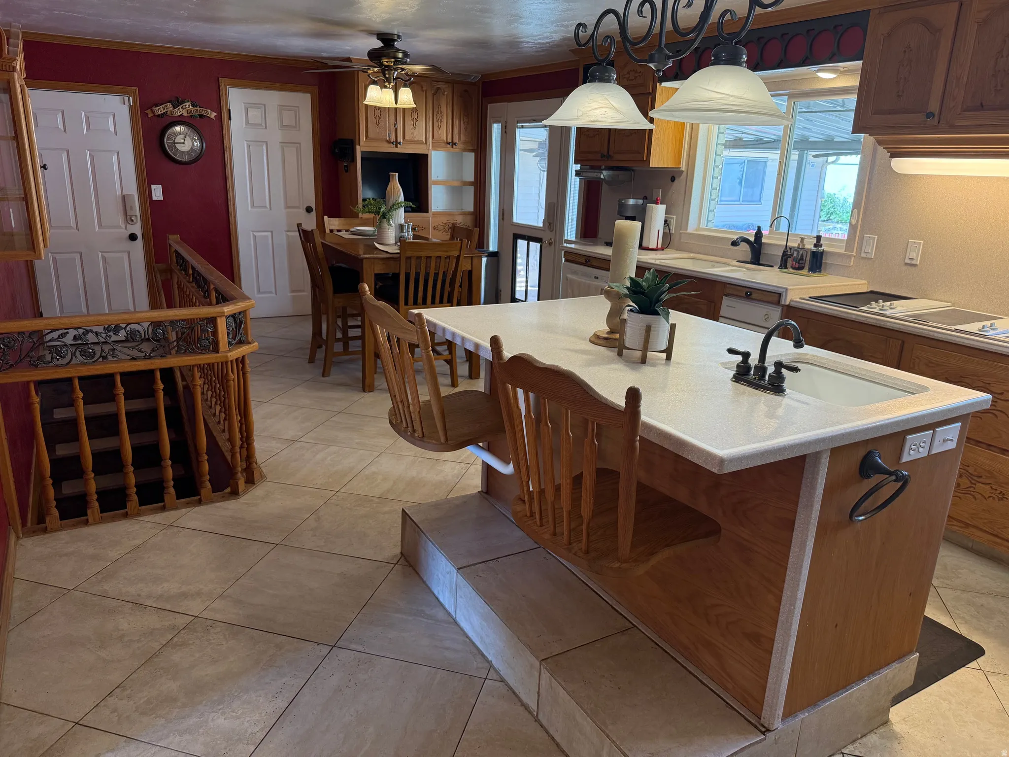 Kitchen island with integrated sink and seating, solid surface countertops, light custom wood cabinetry, tile flooring, and a decorative staircase railing. Additional sink in kitchen.