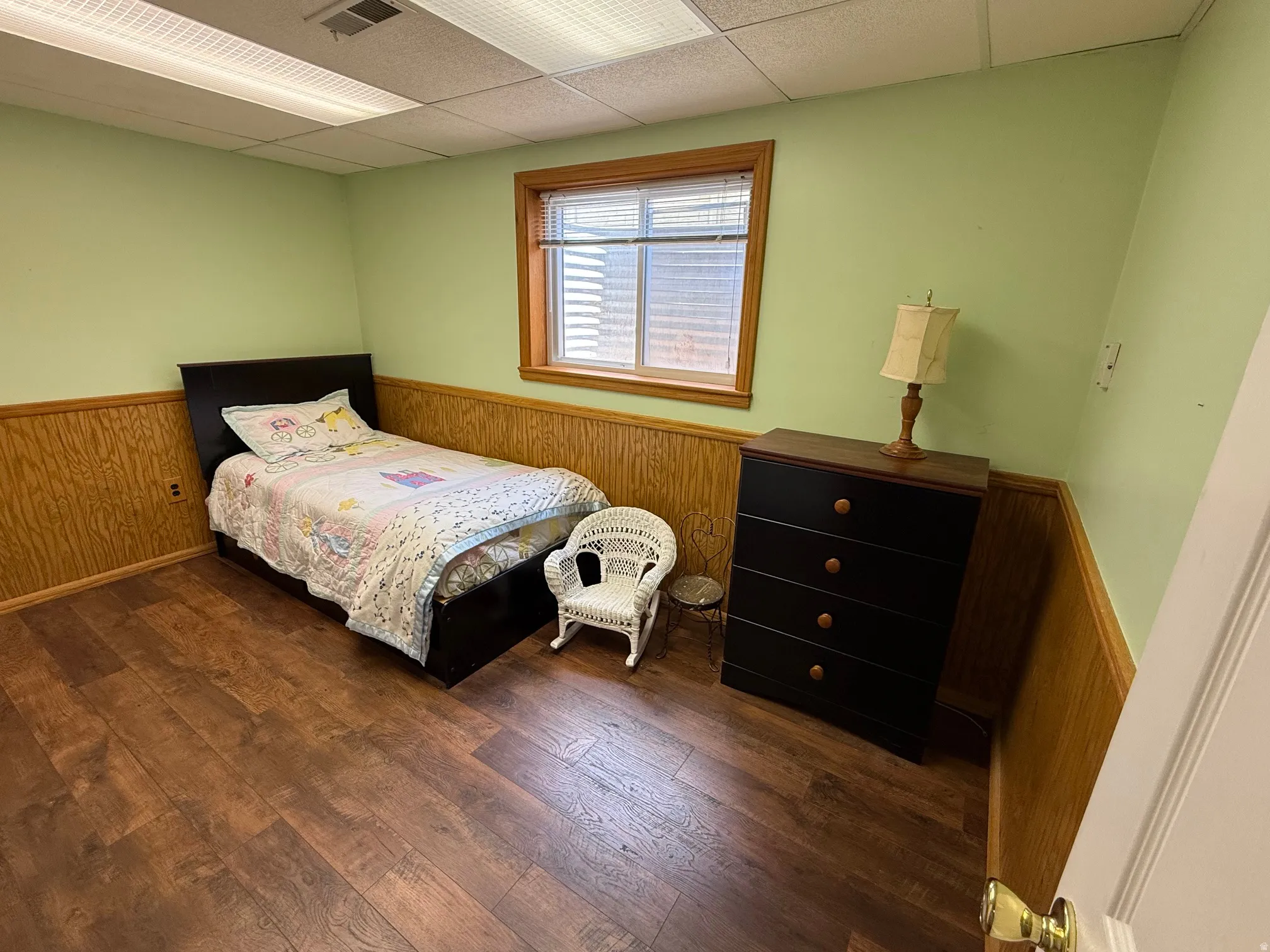 Room featuring wood-finish flooring, half-wall wood paneling, light green wall paint, and a drop ceiling with fluorescent lighting