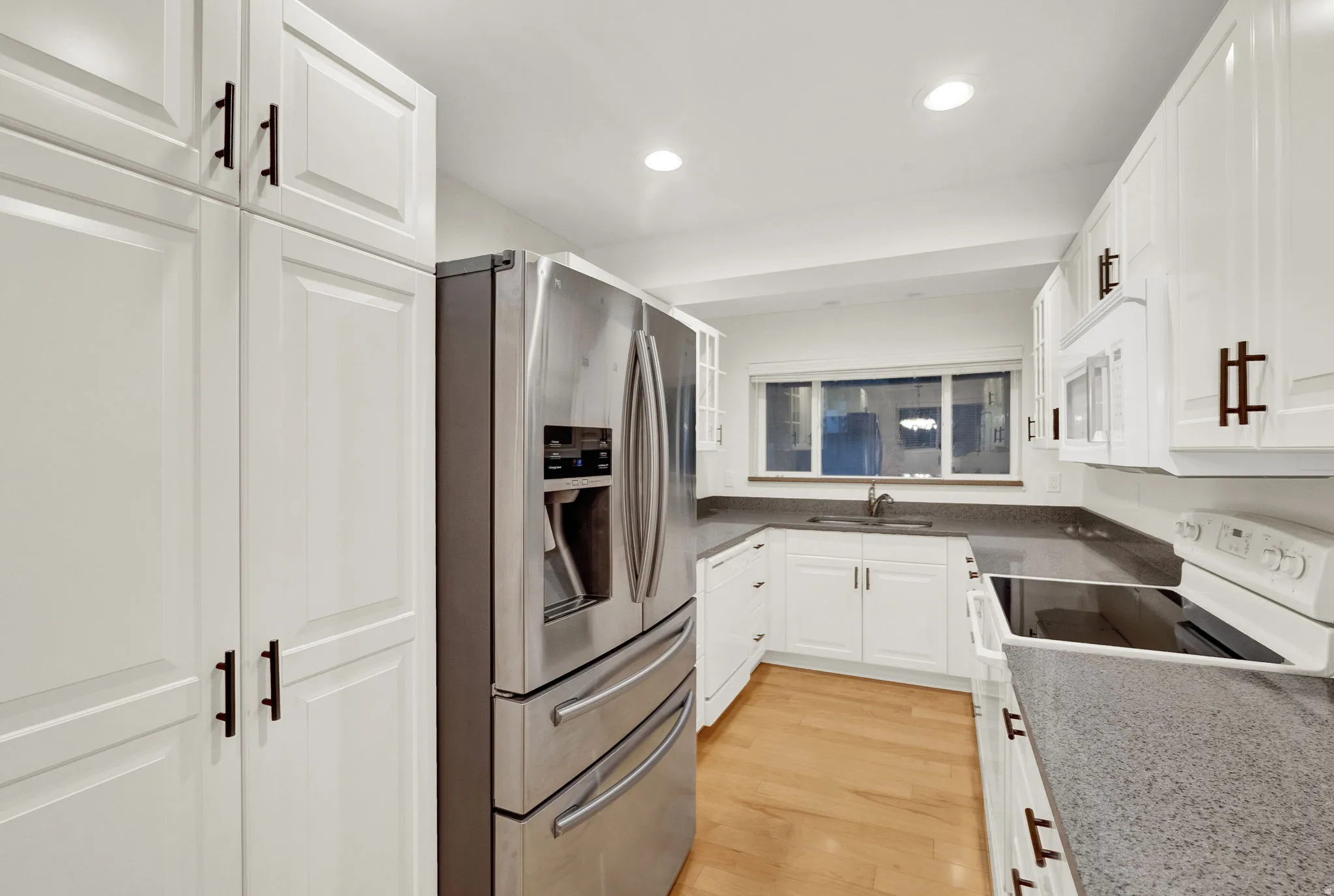 Bright kitchen featuring white shaker-style cabinetry, stainless steel appliances, dark countertops, and wood-finish flooring