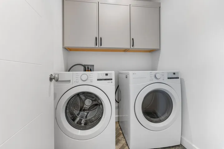 Laundry area featuring stacked front-load machines, light gray upper cabinetry with black hardware, and wood-finish flooring
