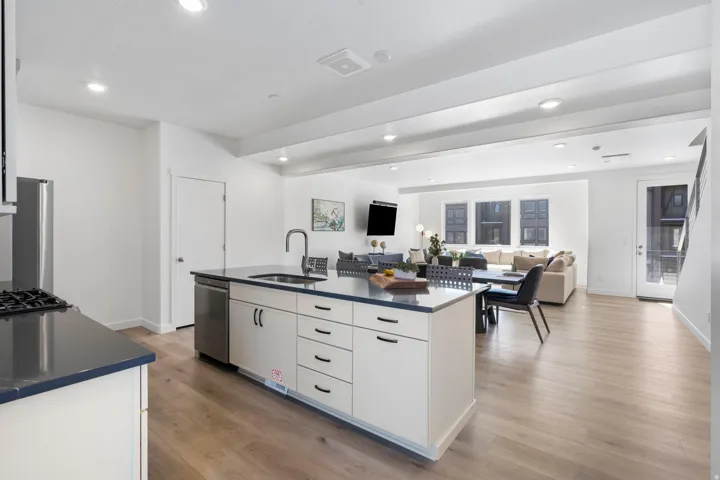 Kitchen island with dark countertop and integrated sink, stainless steel dishwasher, and white cabinetry with dark hardware