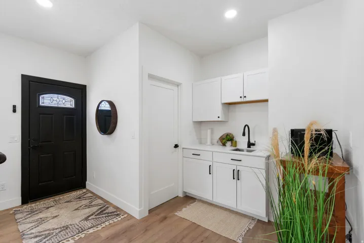 Entryway featuring a black paneled door with decorative glass, wood-finish flooring, white walls, recessed lighting, and a compact kitchenette with white cabinetry, a black sink, and a black faucet