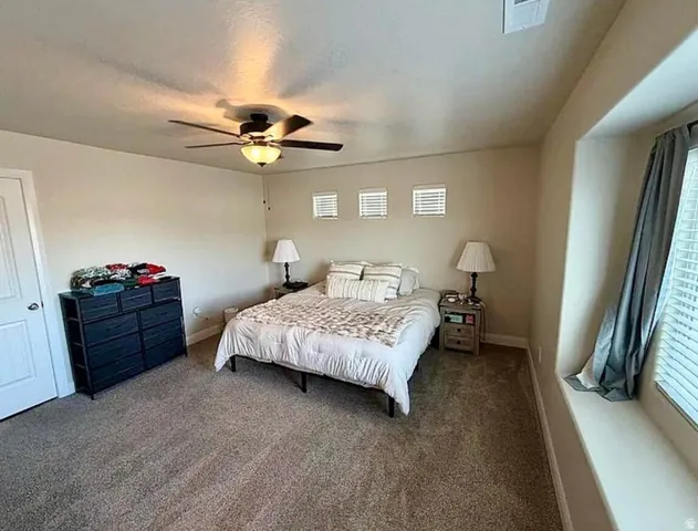 Carpeted bedroom featuring a ceiling fan with integrated lighting, three transom windows, a large window with blinds and drapes, and an architectural window seat