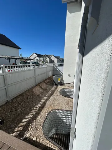 Gravel-lined side yard with a white picket fence, two window wells with grates, a visible AC unit, and a downspout