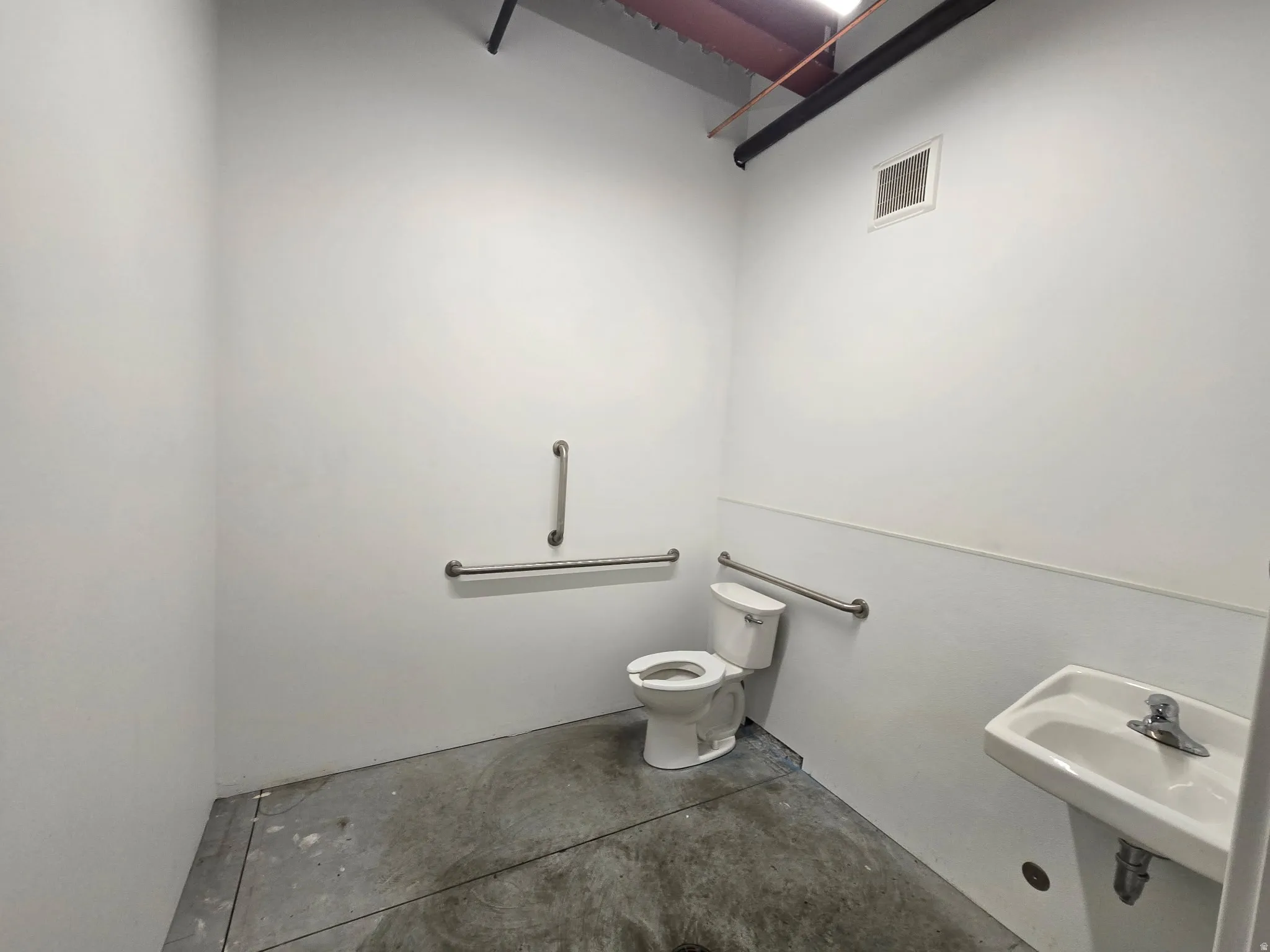 Restroom featuring a wall-mounted sink, a toilet, stainless steel grab bars, and exposed overhead piping