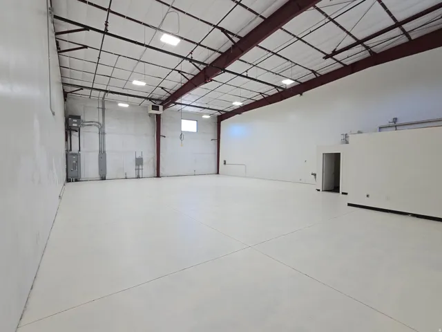 Expansive commercial space featuring a high ceiling with exposed structural beams, white-painted walls, and a light-colored concrete floor