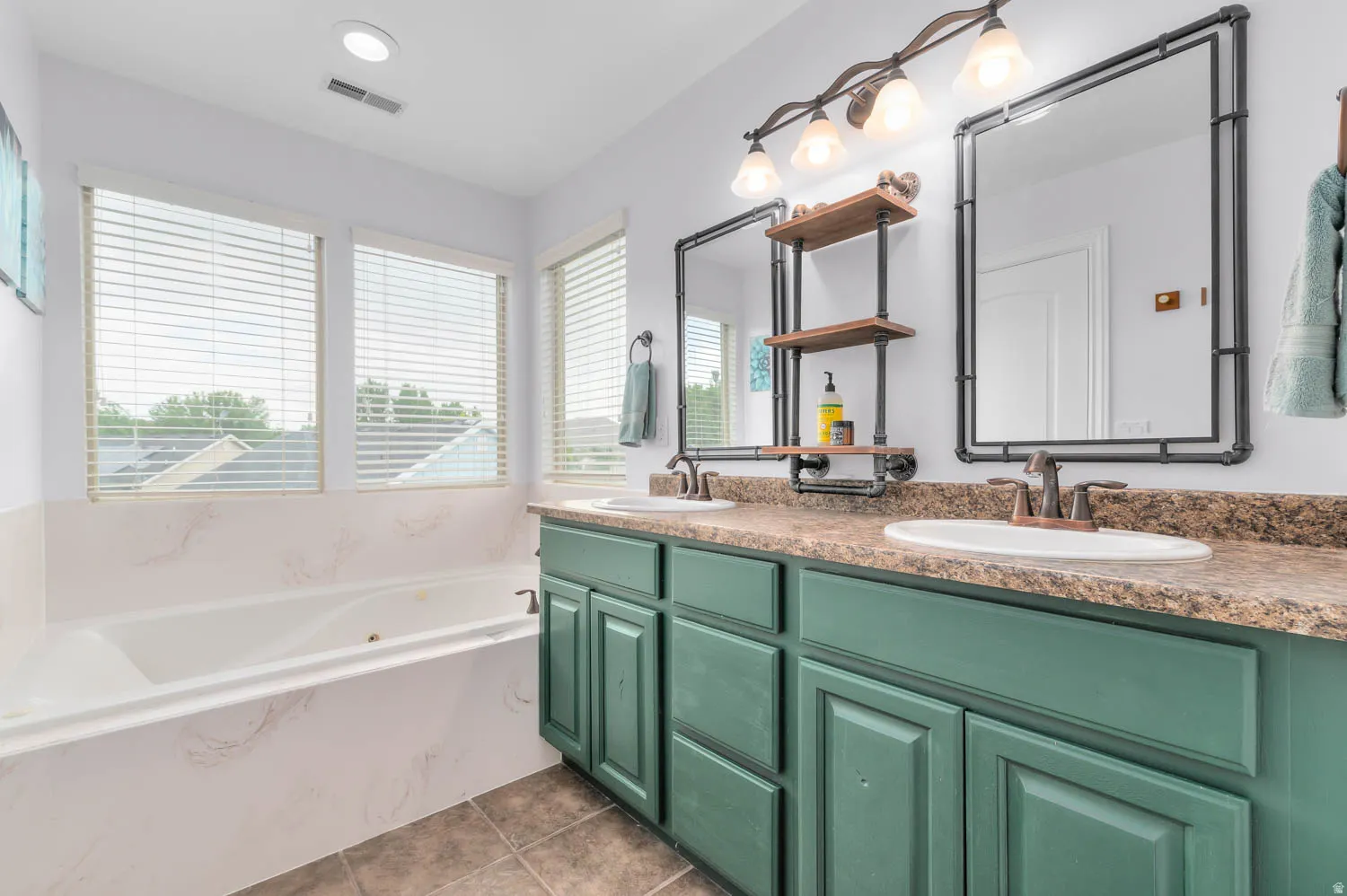 Soaking tub with white surround, three windows with horizontal blinds, dual sink vanity with green cabinetry, wood-finish countertop, and two framed mirrors