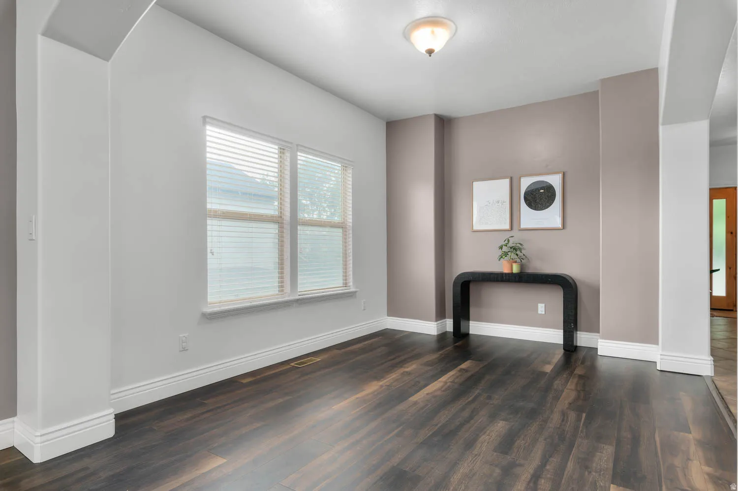 Spacious interior room featuring dark wood-finish flooring, a double window with blinds, and white baseboards