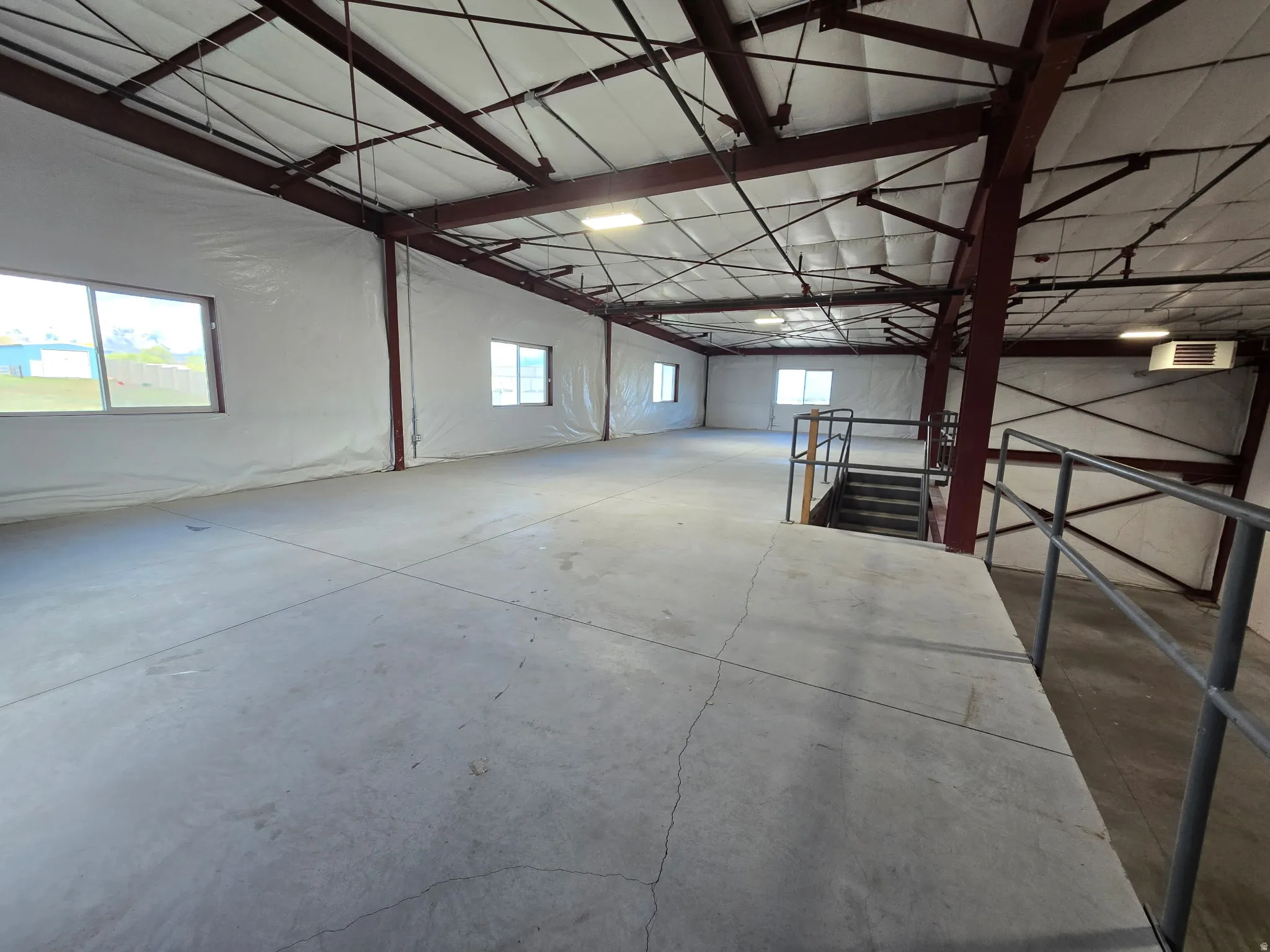Spacious industrial interior featuring exposed steel I-beams, concrete flooring, multiple windows, and a raised platform area with a staircase and handrails