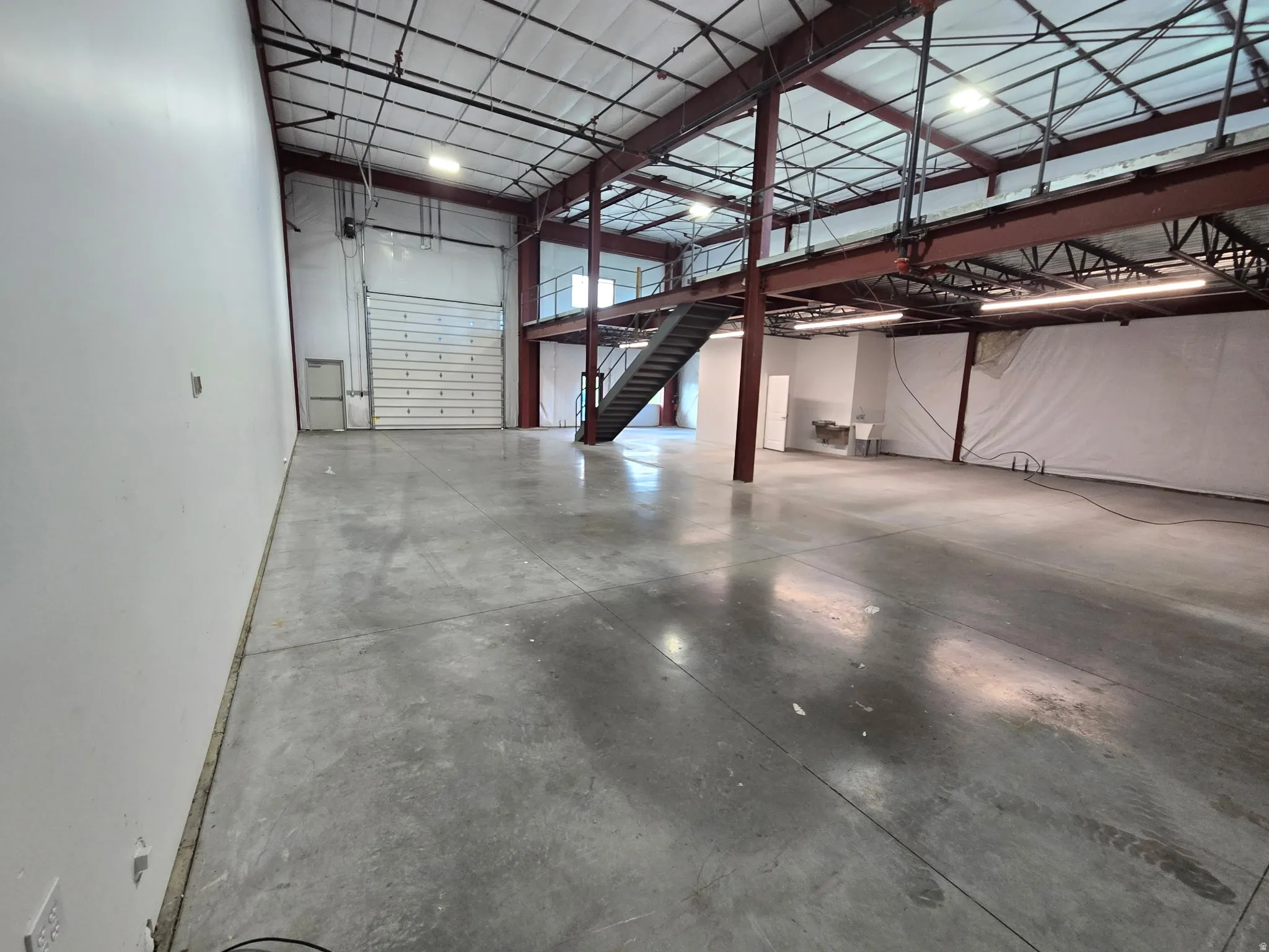 Expansive open warehouse space with polished concrete flooring, exposed red-painted structural beams, and a mezzanine level with staircase access