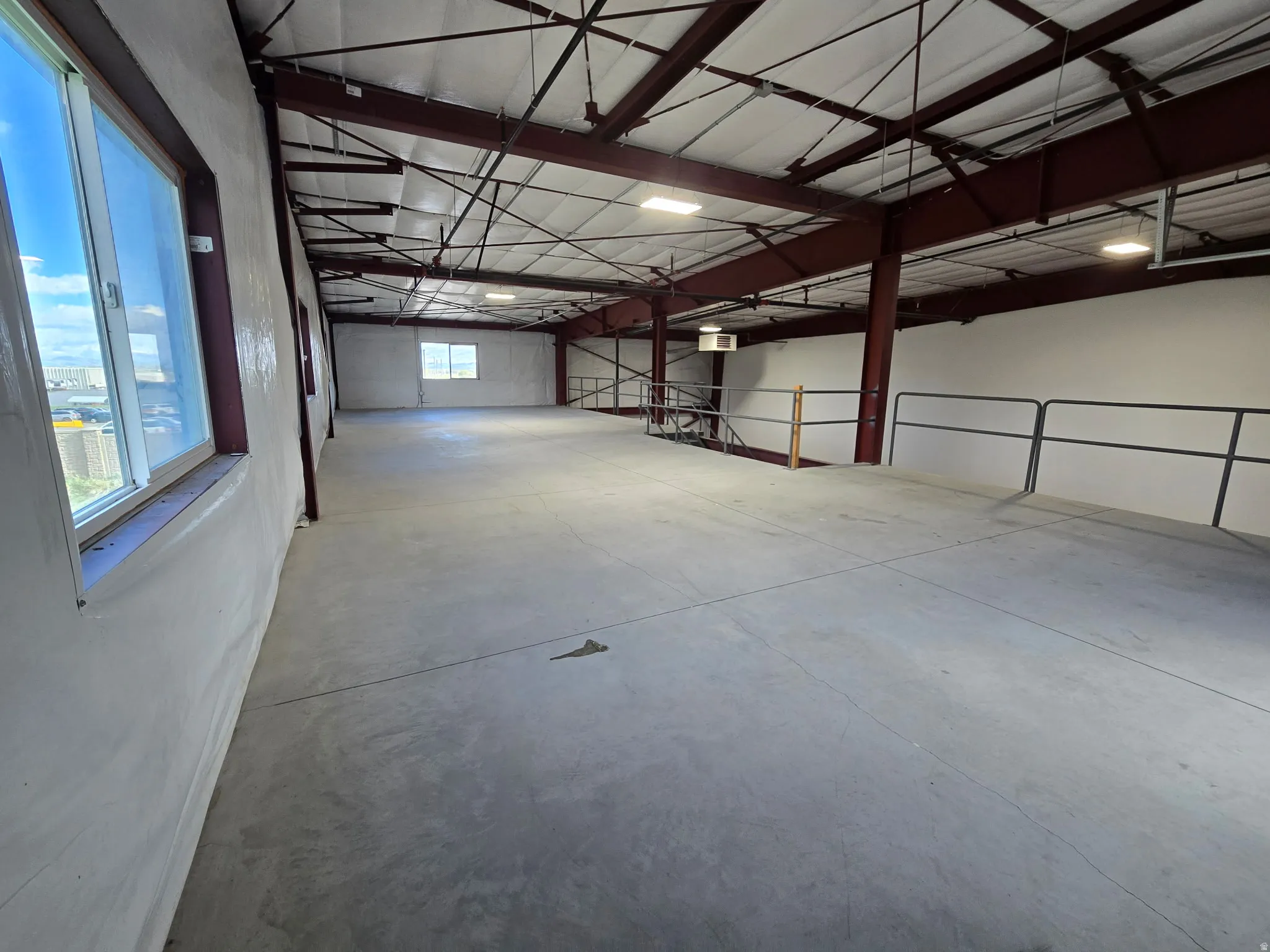 Spacious open-concept commercial interior featuring exposed structural beams, concrete flooring, multiple windows, and a mezzanine level with metal railings