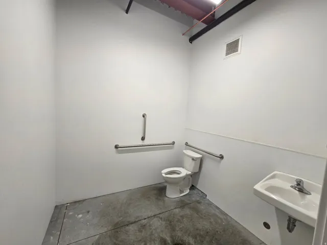 Restroom featuring a wall-mounted sink, a toilet, stainless steel grab bars, and exposed overhead piping