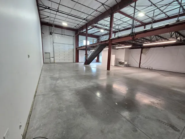 Expansive open warehouse space with polished concrete flooring, exposed red-painted structural beams, and a mezzanine level with staircase access