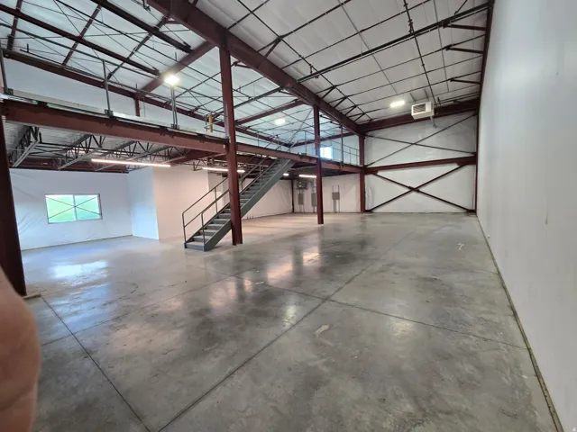 Expansive commercial interior featuring high ceilings with exposed metal trusses, polished concrete flooring, an integrated mezzanine level, a metal staircase with handrails, and multiple wall-mounted light fixtures