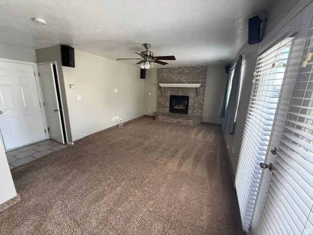 Spacious living area featuring a brick fireplace with a white mantel, ceiling fan, and light tan carpeting