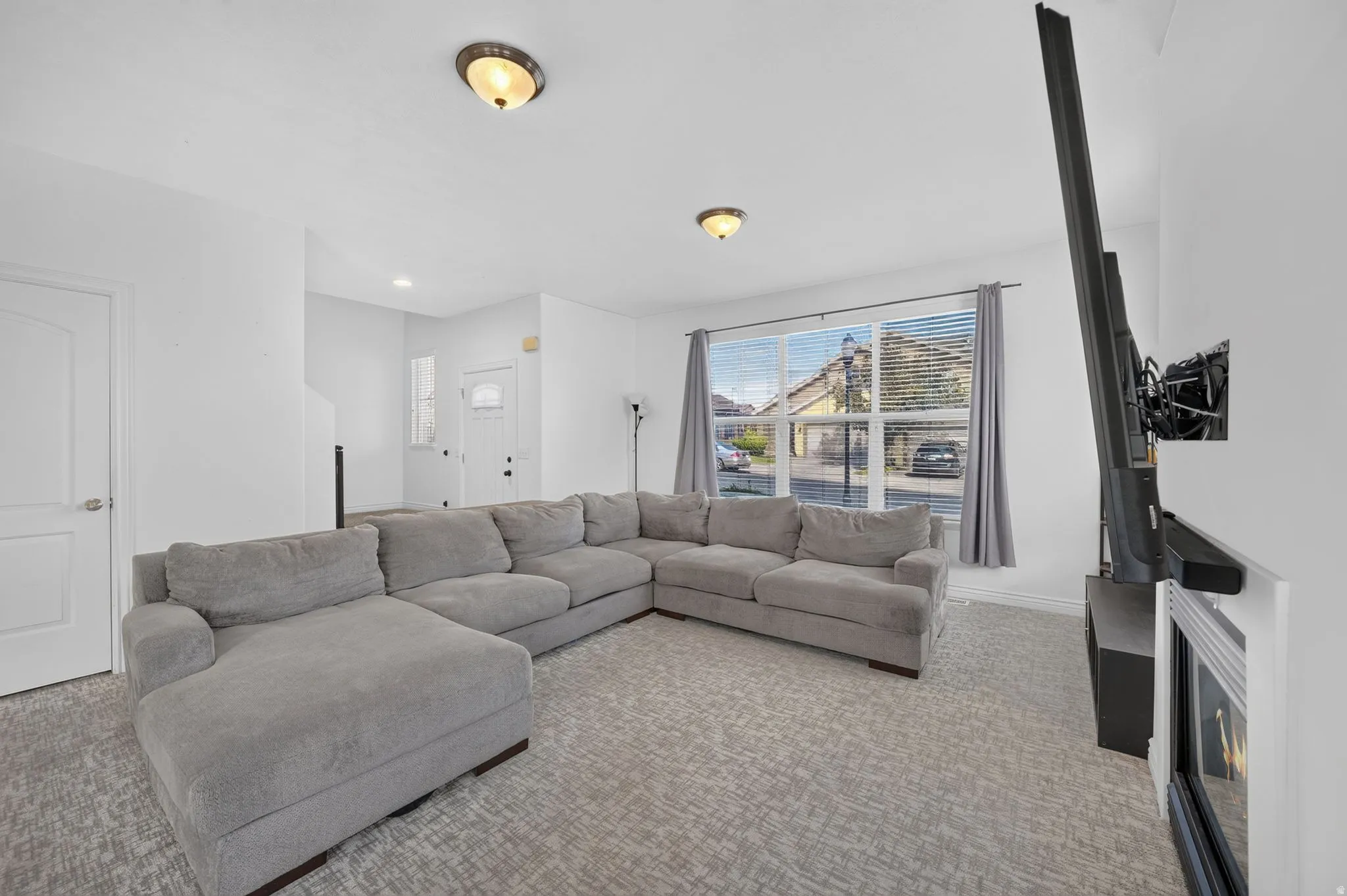 Spacious living area featuring light-toned walls, patterned carpet flooring, and a large window with blinds and drapes