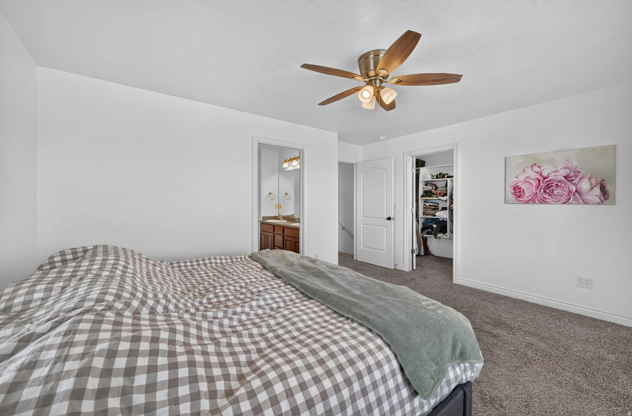 Carpeted bedroom featuring white walls, a ceiling fan with light fixture, and a doorway revealing a bathroom with a wood-finish vanity