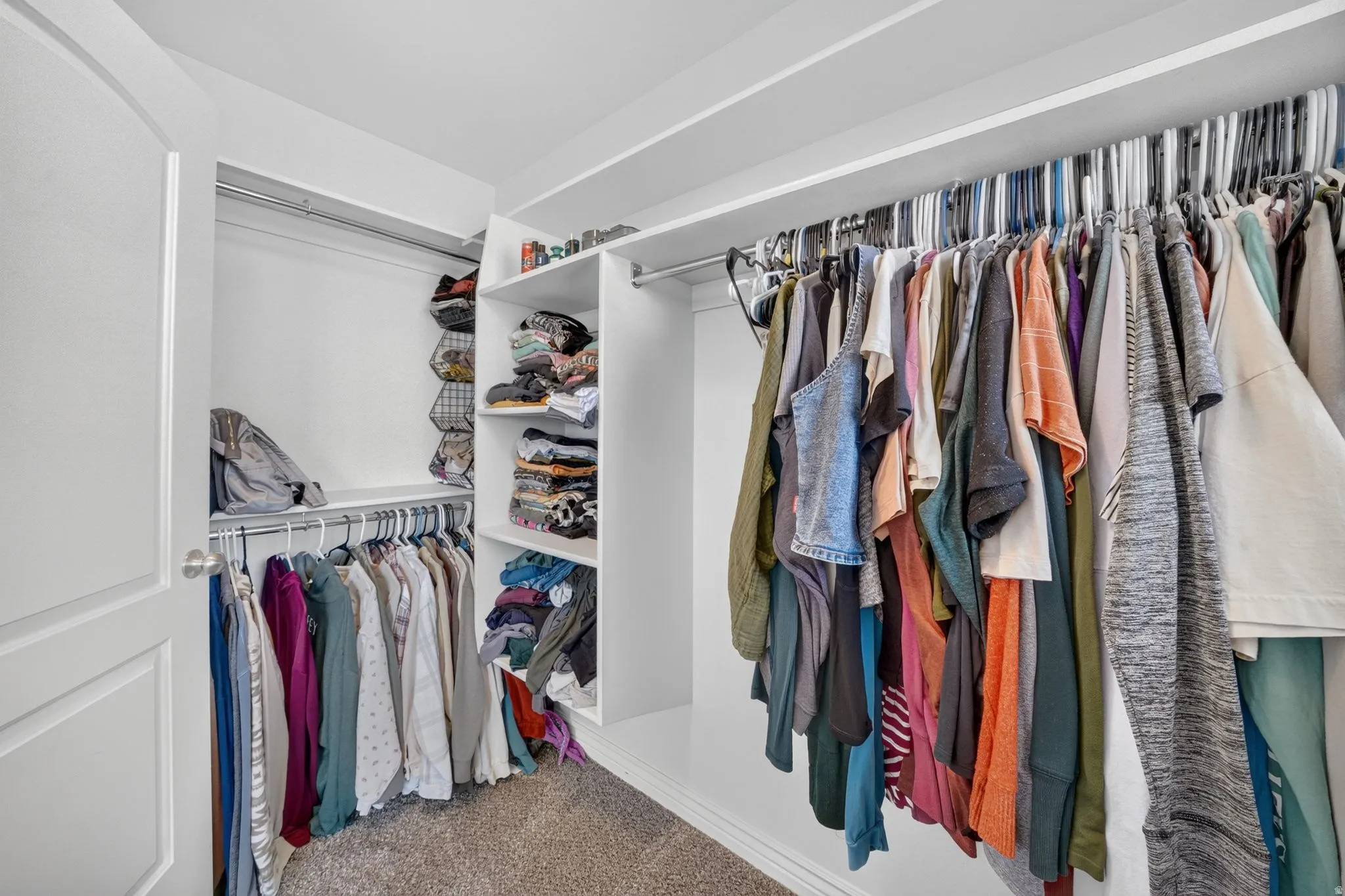 Spacious walk-in closet featuring white built-in shelving, multiple hanging rods, and neutral carpet flooring