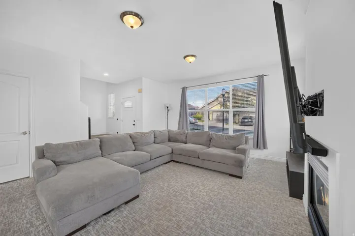 Spacious living area featuring light-toned walls, patterned carpet flooring, and a large window with blinds and drapes