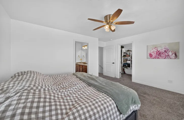 Carpeted bedroom featuring white walls, a ceiling fan with light fixture, and a doorway revealing a bathroom with a wood-finish vanity