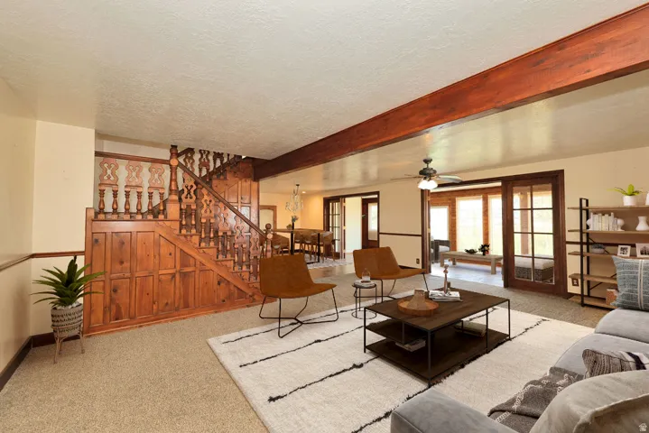 Spacious living area featuring exposed wood beams, neutral carpeting, and a prominent wood staircase with ornate balusters