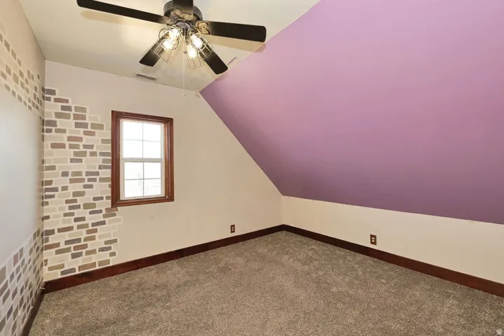 Carpeted room featuring a ceiling fan with integrated lighting, a window with wood trim, and a sloped ceiling