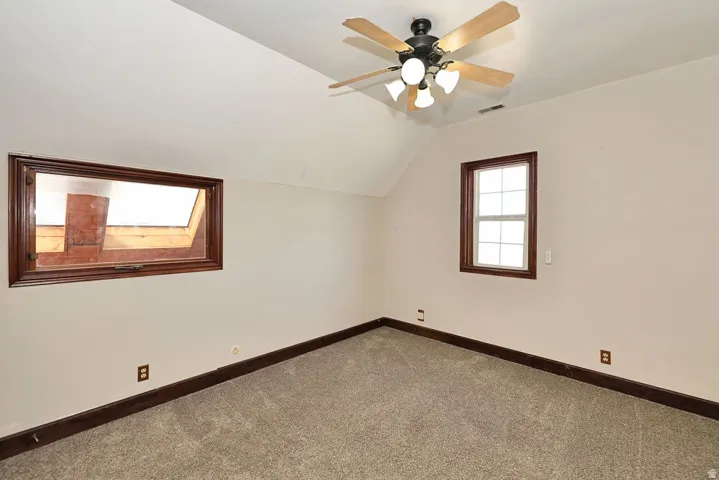 Carpeted room featuring a sloped ceiling, a ceiling fan with integrated lighting, and two windows with dark wood trim
