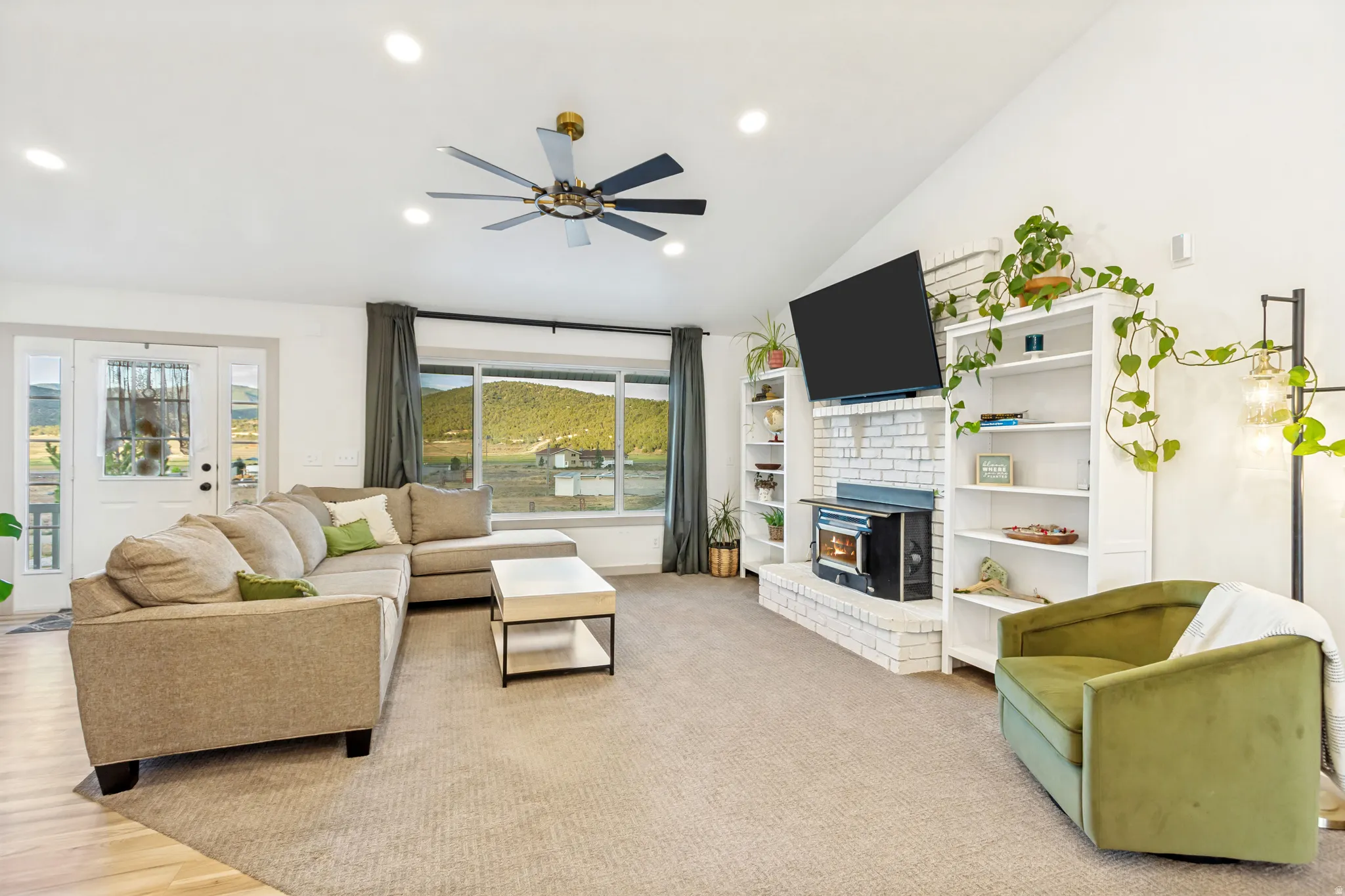 Vaulted ceiling living area featuring recessed lighting, a modern ceiling fan, a white painted brick fireplace with a wood-burning stove, built-in shelving, and large windows