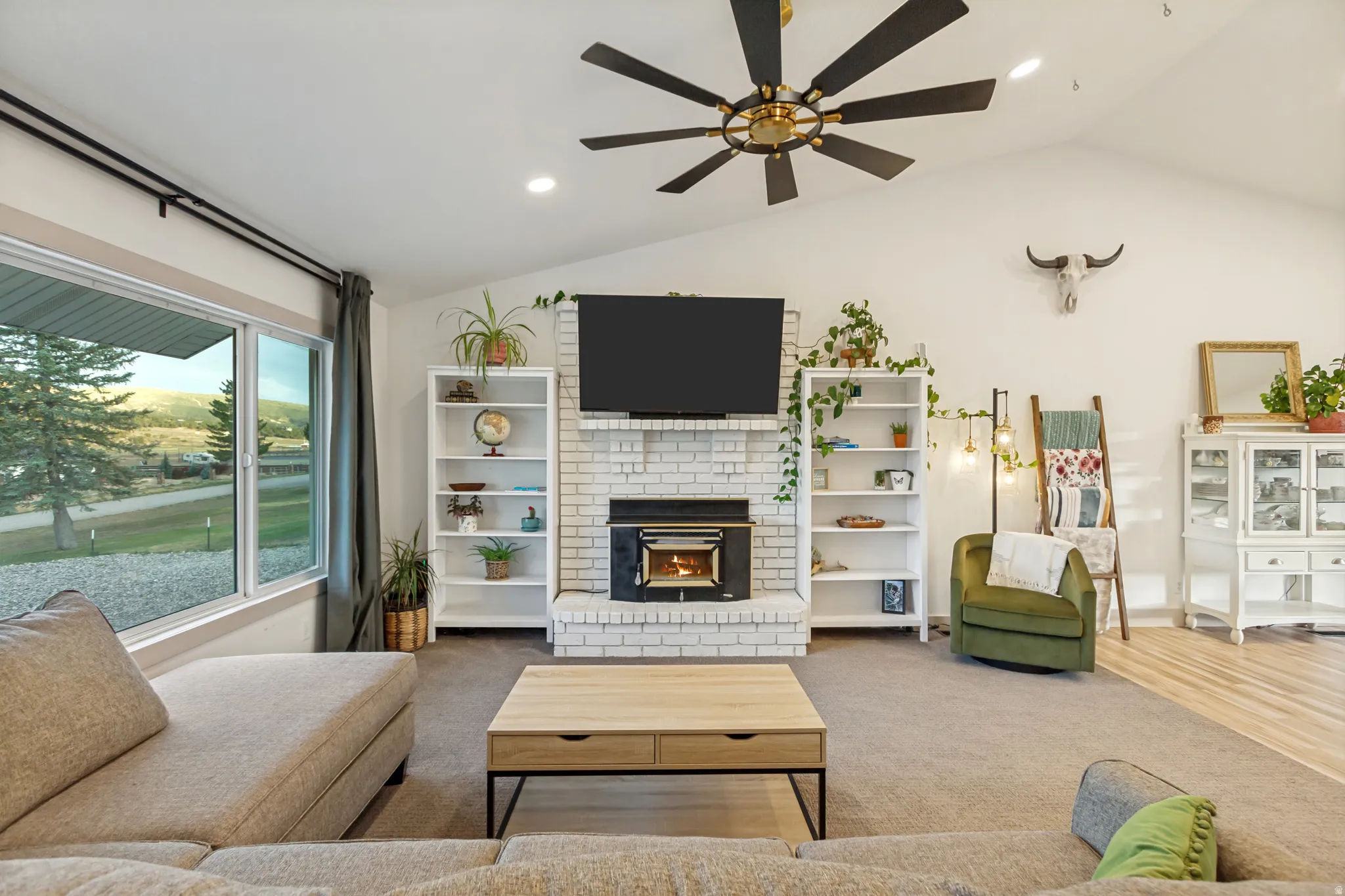 Living area featuring a vaulted ceiling with a large ceiling fan, white brick fireplace with an insert, built-in shelving, expansive windows, and light wood-finish flooring