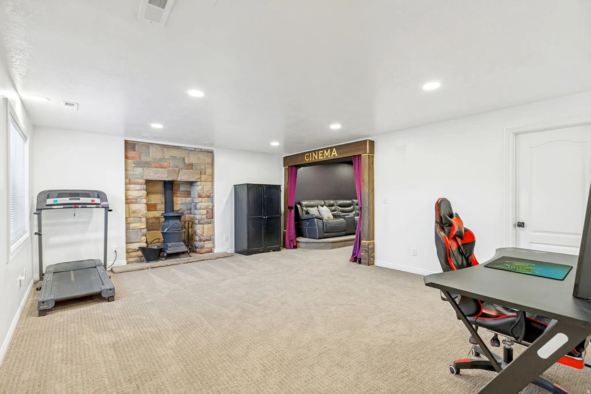 Expansive carpeted room featuring a stone-clad hearth with a black wood stove, recessed lighting, and a dedicated home cinema entrance with purple drapes