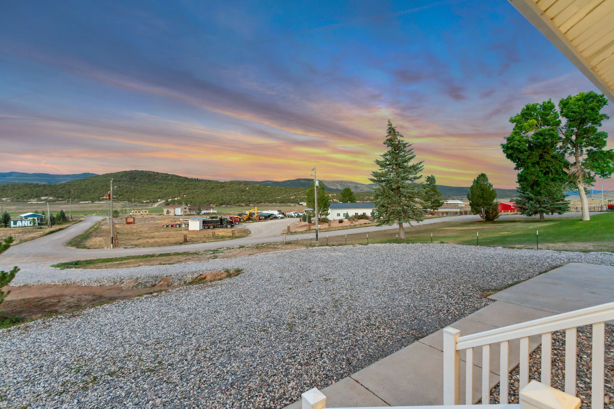 Gravel driveway with concrete walkway and white railing, framed by mature trees and distant mountains under a colorful sky