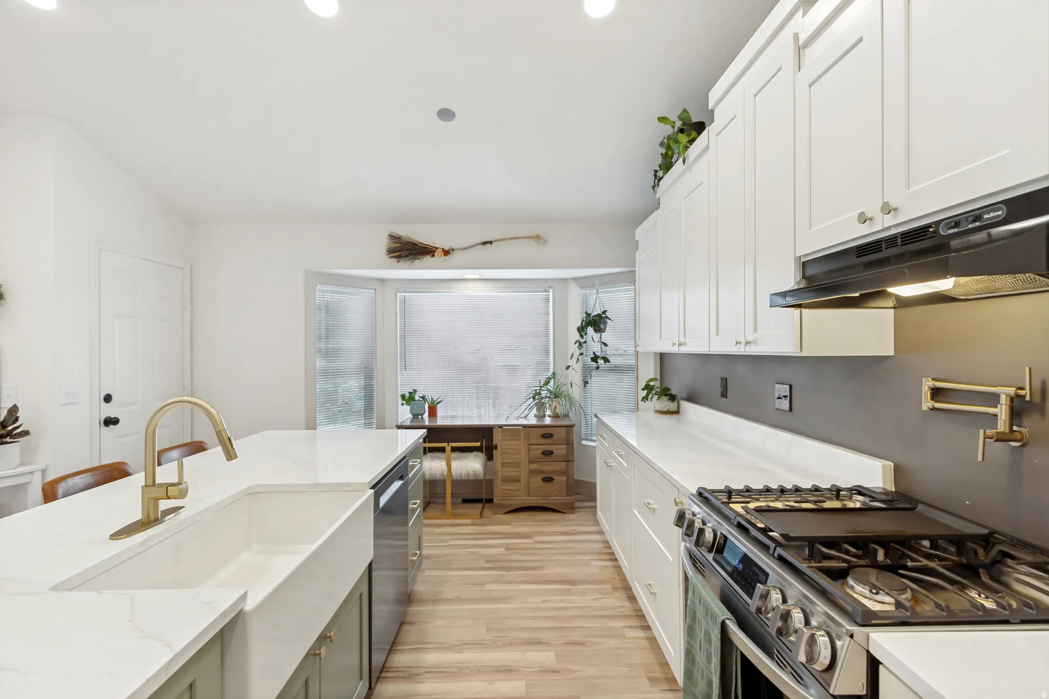 Contemporary kitchen featuring white shaker cabinetry, white countertops, wood-finish flooring, a farmhouse sink with a gold-tone faucet, and stainless steel appliances
