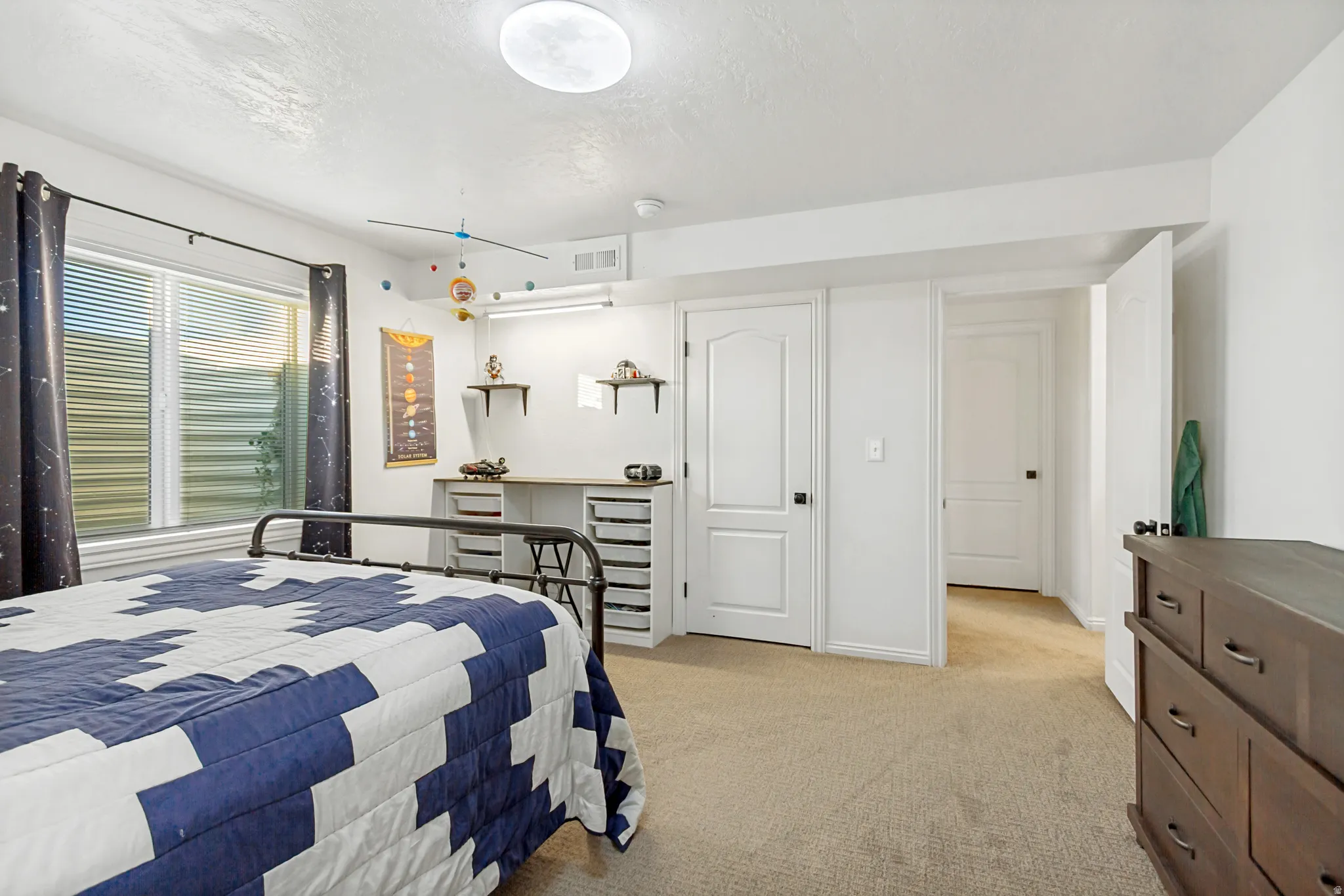 Carpeted bedroom featuring a large window with blinds, a built-in desk area with shelving, and two panel doors