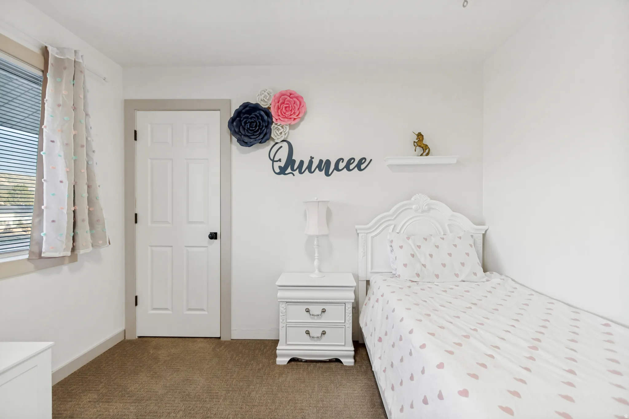 Crisp white walls frame a room featuring a rectangular window with blinds, a white paneled door with dark hardware, and plush carpet flooring