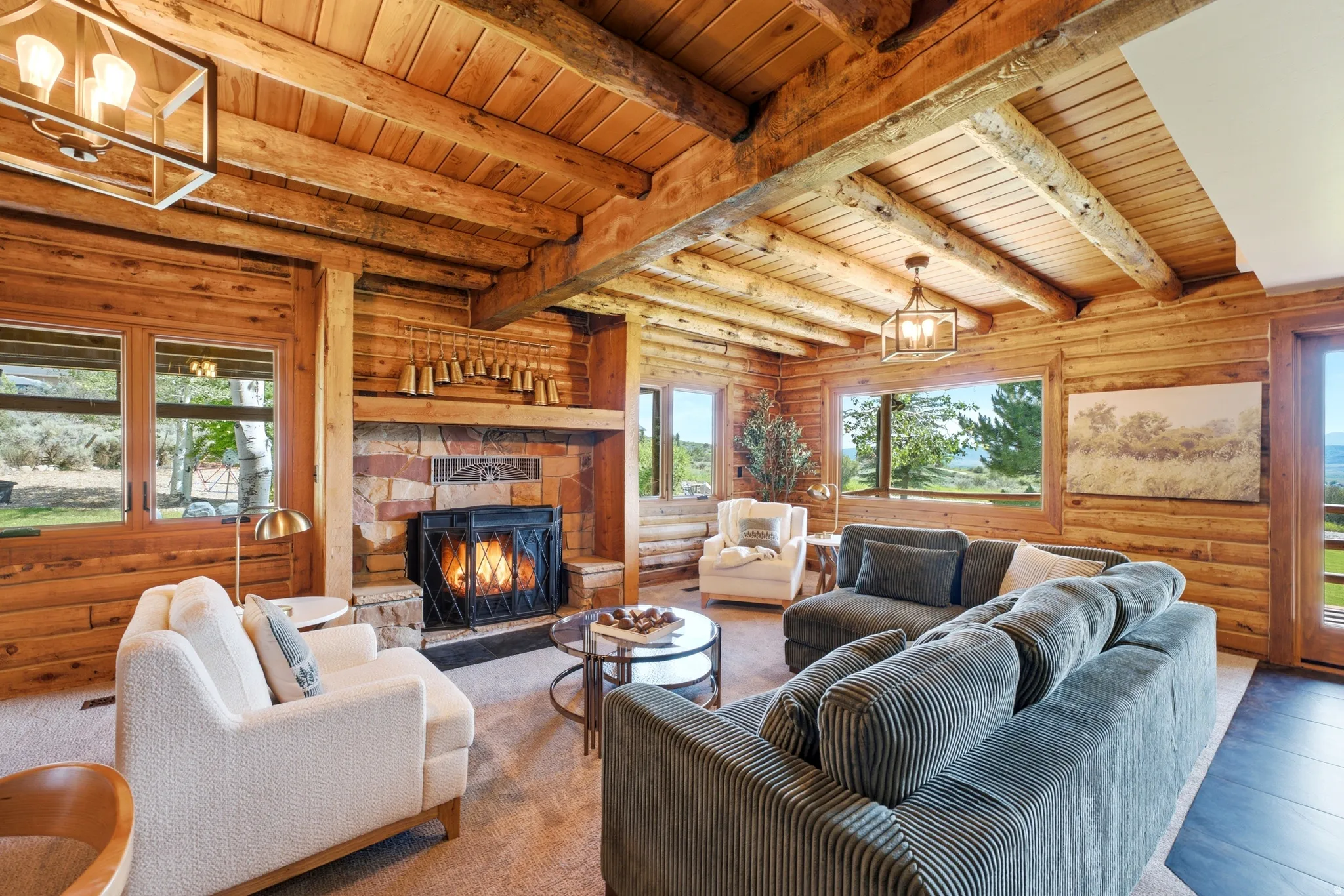 Living area featuring exposed log walls and ceiling beams, a stone-surround fireplace with a metal grate, large windows, wood-finish flooring, and a glass-paneled door