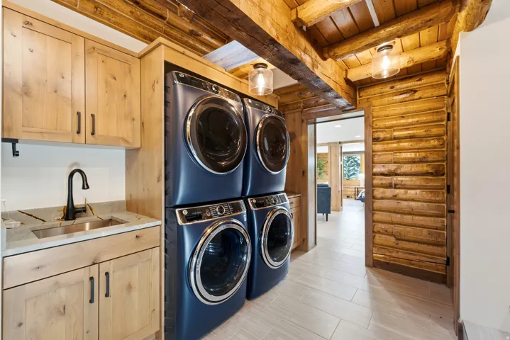Laundry area featuring light wood cabinetry with dark hardware, a stone-finish countertop with an undermount sink and matte black faucet, and stacked front-load washers and dryers