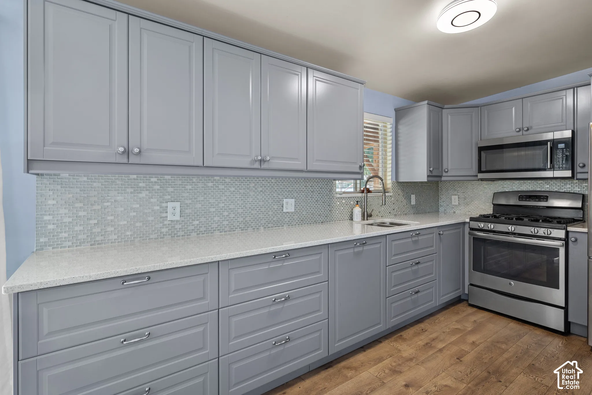 Kitchen featuring gray shaker-style cabinetry, solid-surface countertops, mosaic tile backsplash, stainless steel appliances, and wood-finish flooring