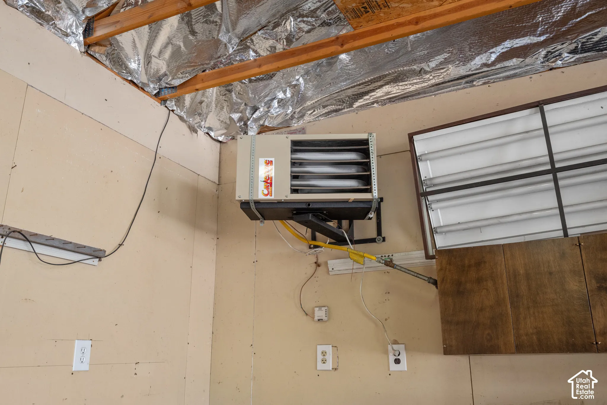 Wall-mounted forced-air heater with exposed framing and reflective insulation, including overhead fluorescent lighting fixtures and wall-mounted electrical outlets