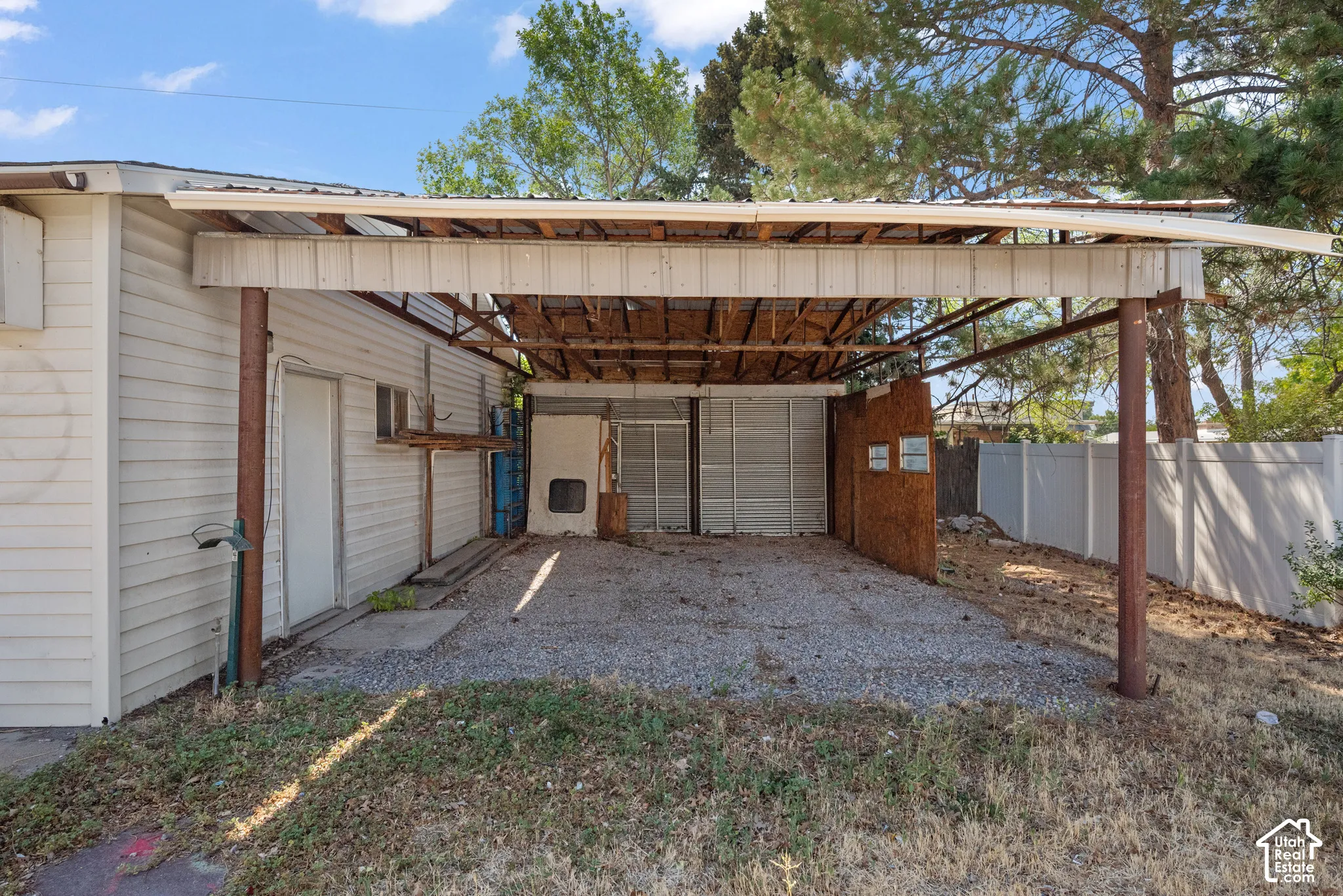 Covered carport with a gravel surface, metal support columns, and white privacy fencing