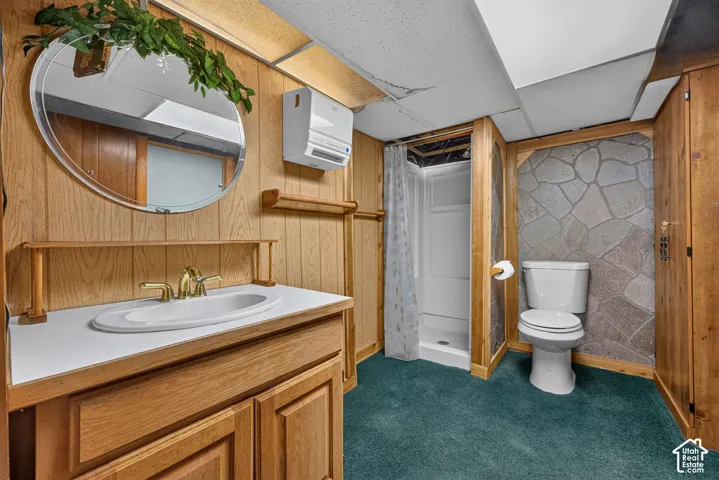 Bathroom featuring wood-paneled walls, an oval mirror, a vanity with a white countertop and gold-finish faucet, an enclosed shower, and a stone-accent wall
