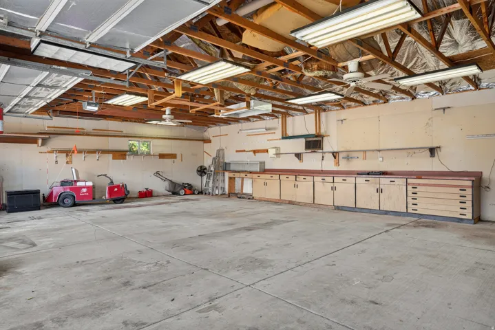 Spacious garage featuring a concrete floor, exposed wood beam ceiling, multiple fluorescent light fixtures, a window providing natural light, and extensive built-in cabinetry with integrated drawers and countertops