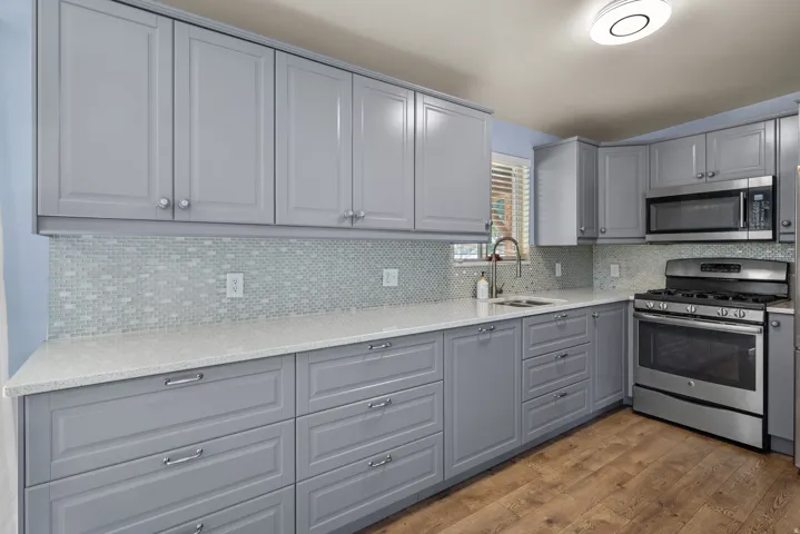 Modern kitchen featuring gray shaker-style cabinetry, light-toned countertops, a full mosaic tile backsplash, stainless steel appliances, and wood-finish flooring