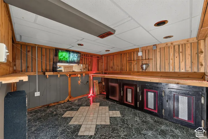 Custom wet bar featuring wood-panelled walls, built-in lower cabinetry with metal-finish inlays, stone-tile flooring with decorative inlay, and a drop ceiling