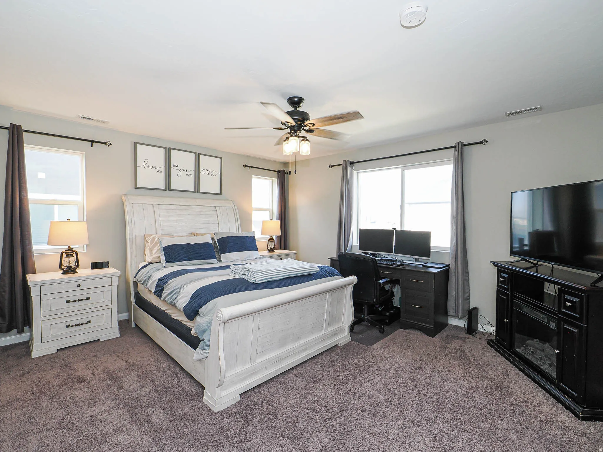 Carpeted room featuring a ceiling fan with light fixture, multiple windows with dark curtain rods, and a built-in electric fireplace media console