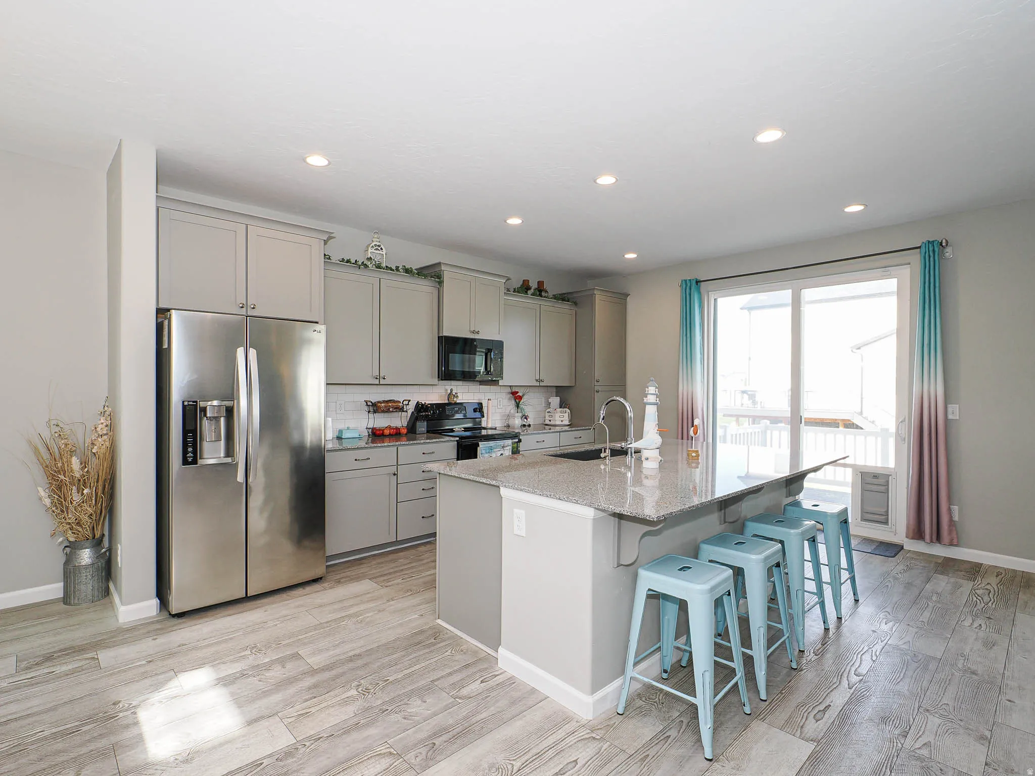 Bright kitchen featuring light gray cabinetry, stainless steel appliances, a large center island with a sink and seating, wood-finish flooring, and a sliding glass door
