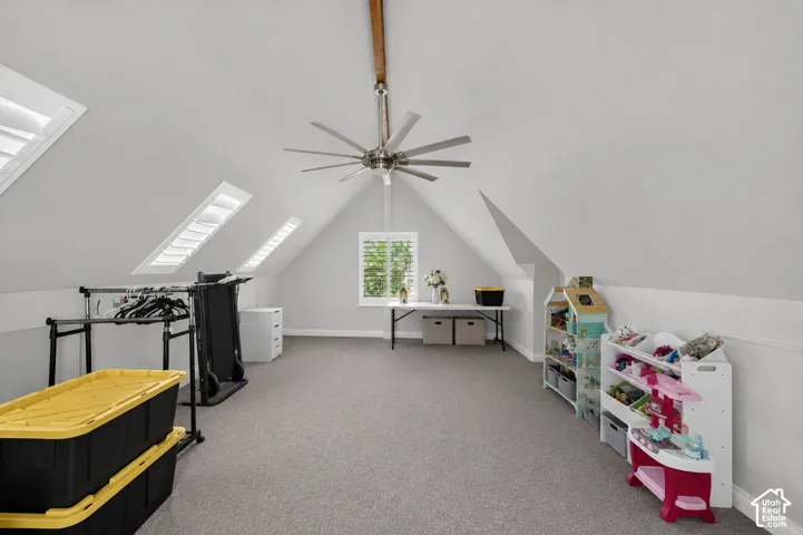 Spacious upper-level room featuring angled ceilings, gray carpeting, a window with plantation shutters, multiple skylight windows, and a ceiling fan with integrated lighting