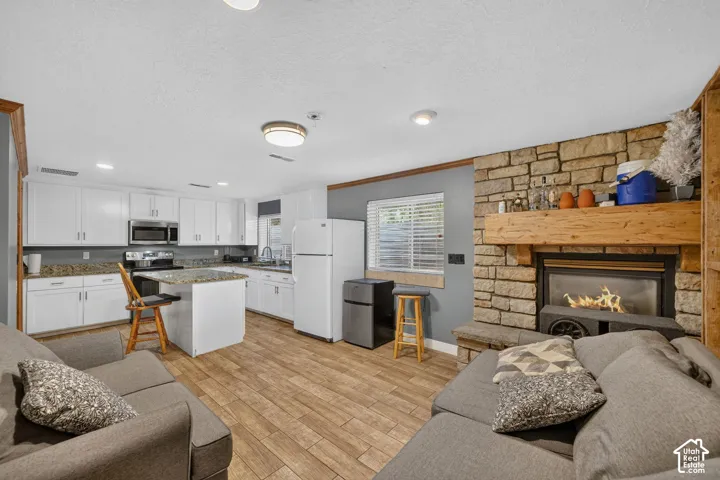 Open-concept living area featuring wood-finish flooring, a stone-faced fireplace with a substantial wood mantel, white cabinetry, granite countertops, and stainless steel kitchen appliances