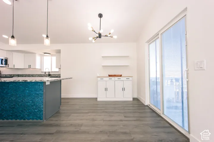 Open-concept space featuring wood-finish flooring, a kitchen island with patterned stone surfacing, minimalist multi-bulb pendant lighting, built-in cabinetry with wall-mounted floating shelves, and sliding glass doors